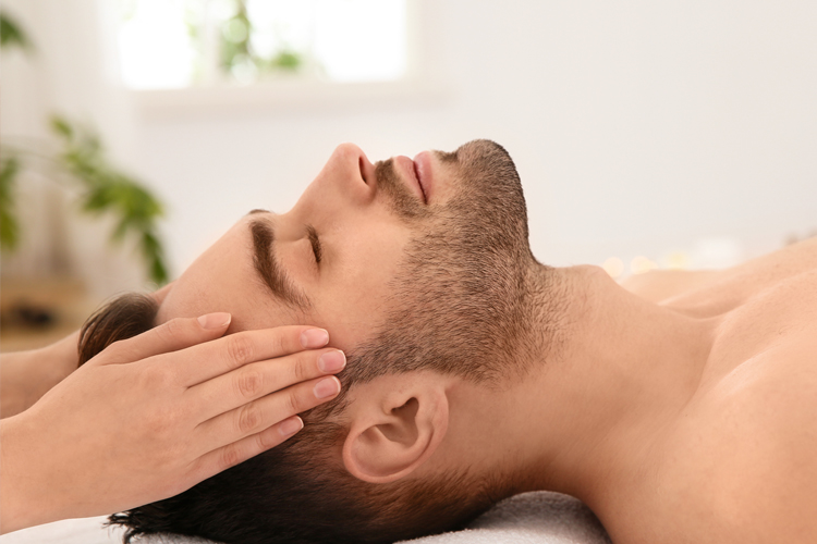 Massagen / Manuelle Therapien, Bild eines jungen Herrn, der entspannt auf dem Rücken liegt. An seinem Kopf befinden sich zwei weibliche Hände, die seinen Kopf massieren.